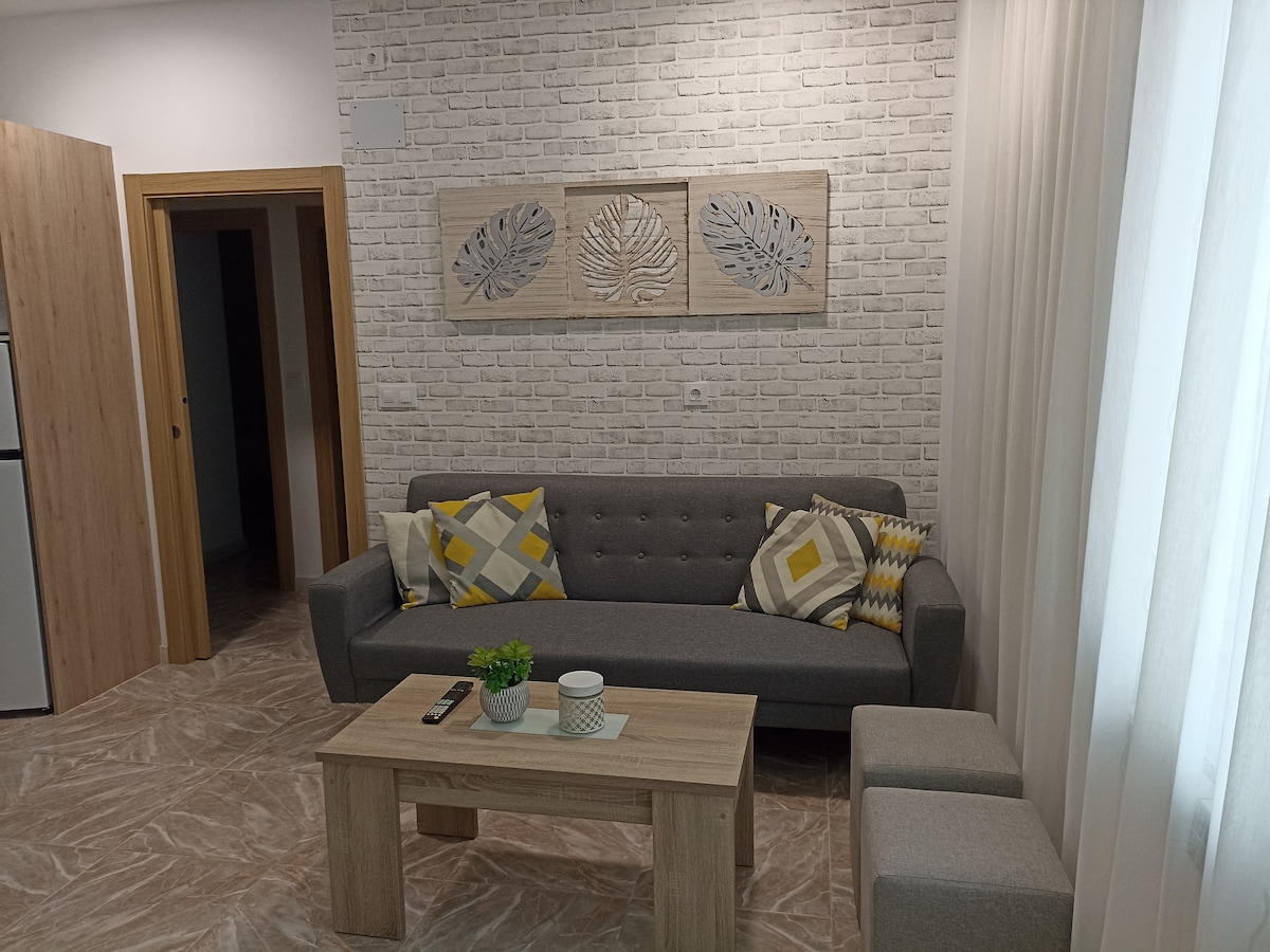 A modern living area features a gray sofa adorned with yellow and white accent pillows. A light wooden coffee table sits in front, complemented by two small stools. The walls are highlighted by a decorative wooden panel, and sheer curtains soften the natural light filtering through.
