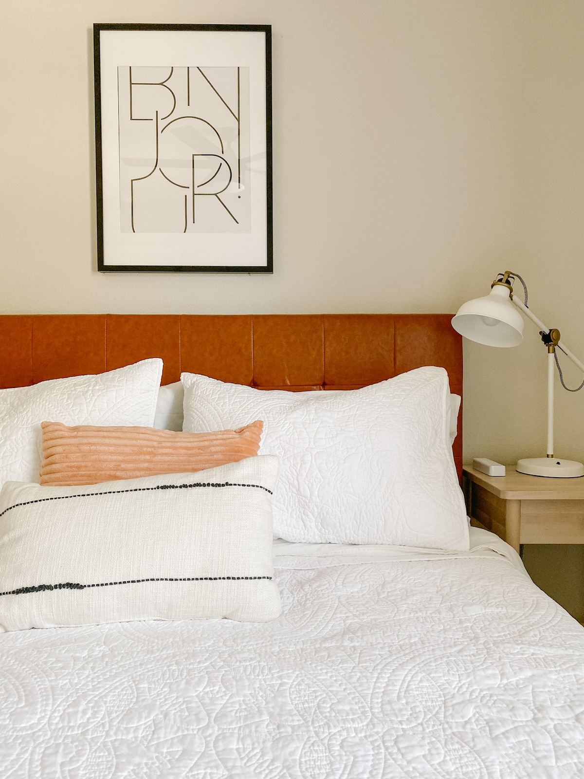 A queen bed is styled with white bedding and decorative pillows, accompanied by a bedside table and lamp. A piece of artwork featuring minimalist typography is hung above the headboard, contributing to the room's aesthetic.
