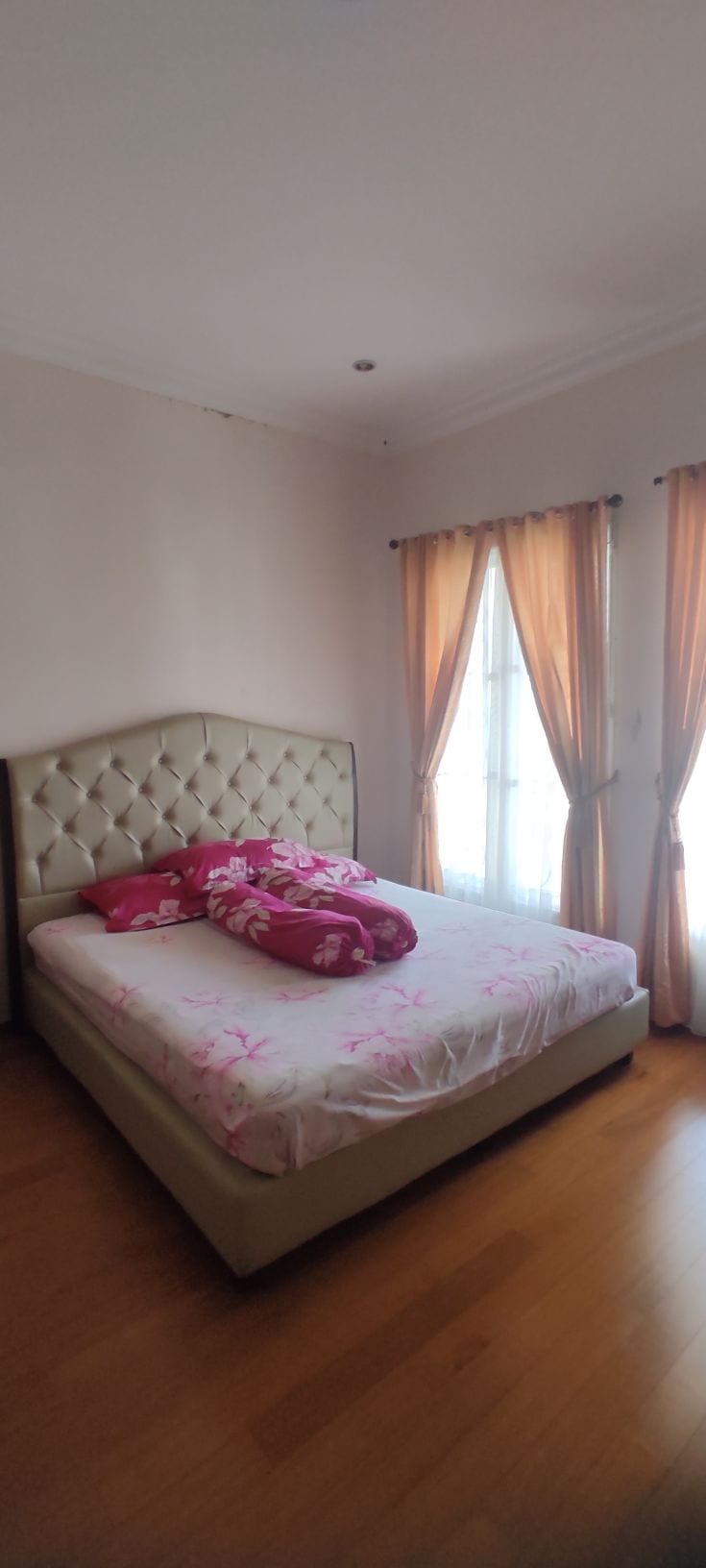 A well-lit bedroom features a headboard with a diamond pattern and a light-toned bed frame. The bed is dressed with a floral-patterned blanket and additional pillows. Two large windows with light, flowing curtains allow natural light to filter in, enhancing the welcoming ambiance.