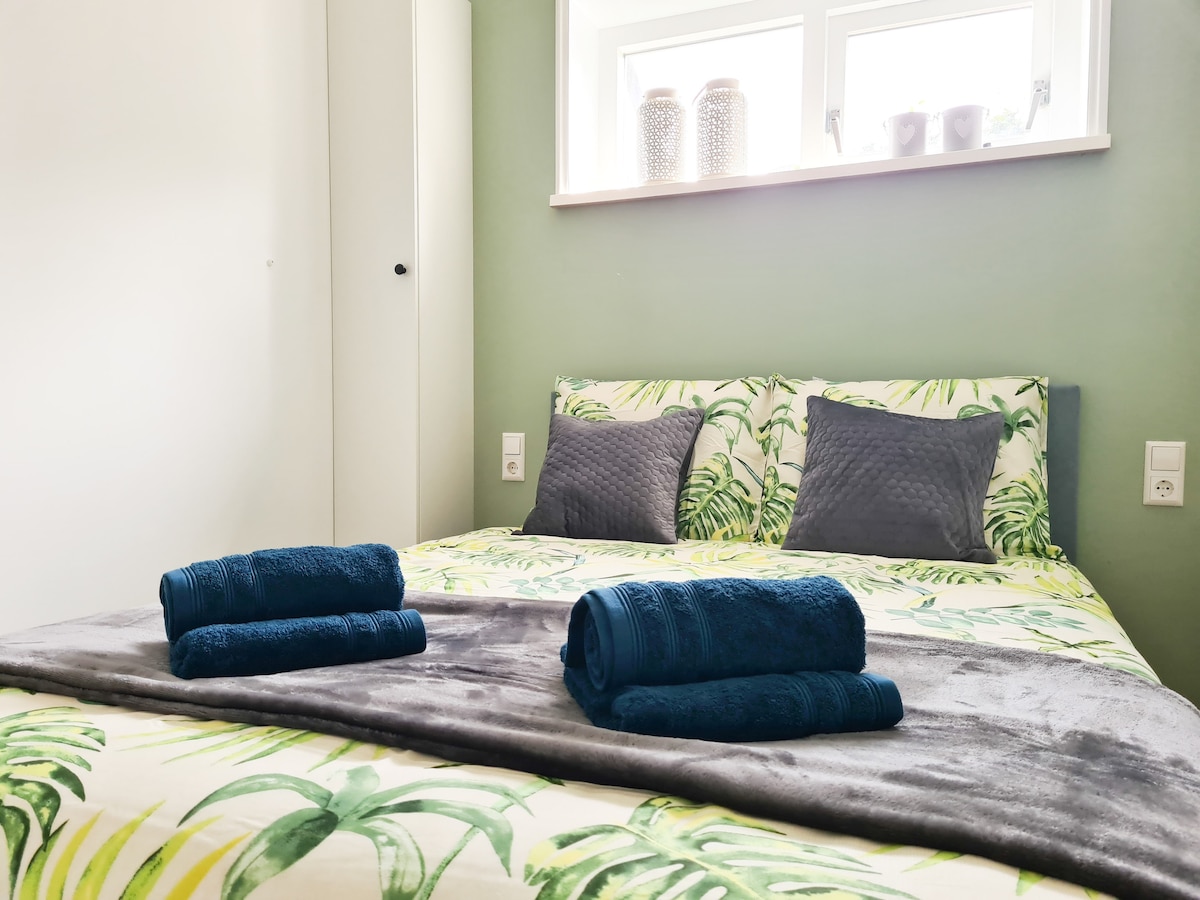 A neatly arranged sleeping area features a bed adorned with leaf-patterned bedding. Dark towels are rolled neatly on the bed, and soft pillows offer comfort. Natural light enters through a window above the bed, which is surrounded by simple decor.