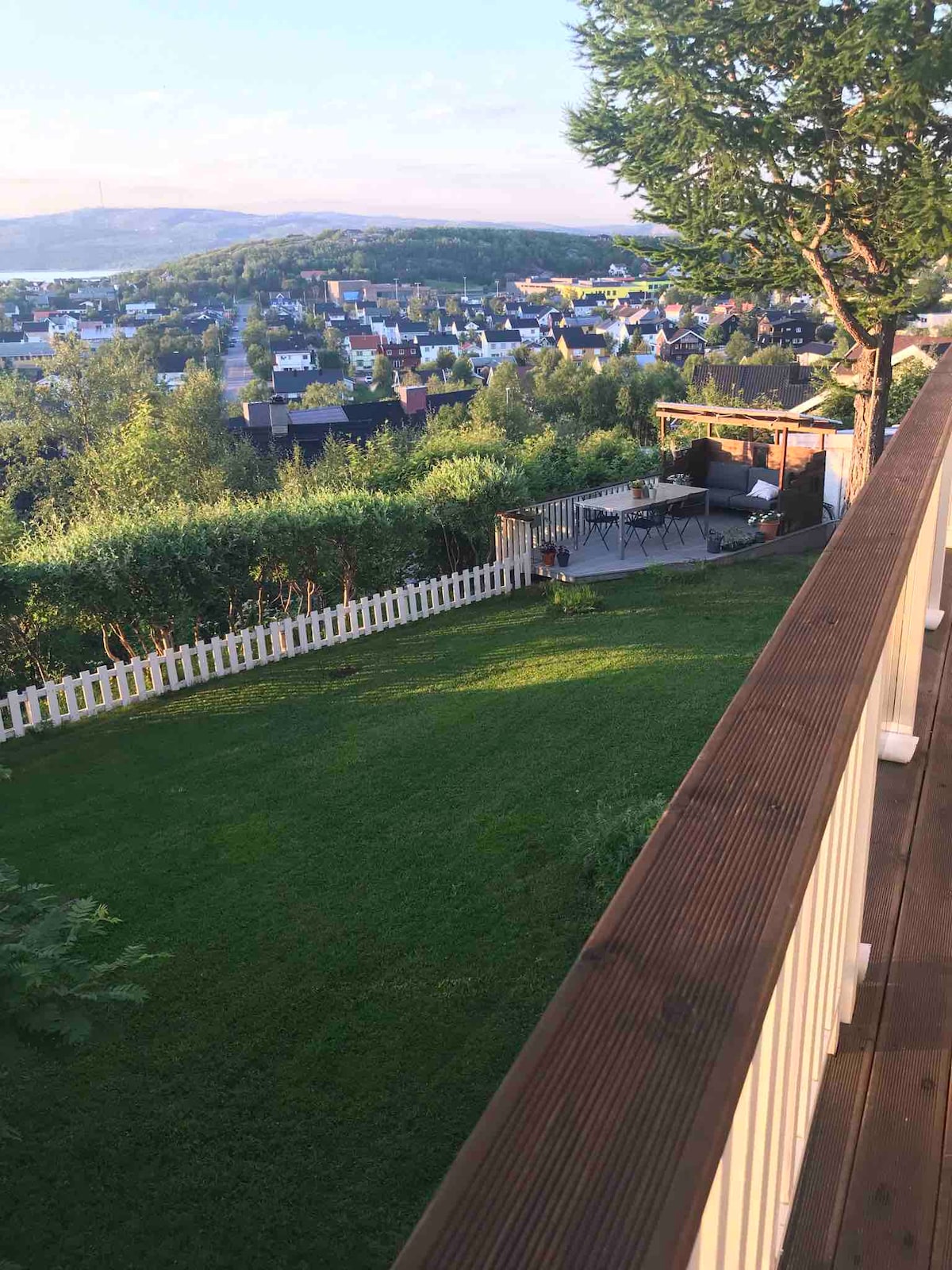 A spacious garden area leads to a wooden deck with a railing, overlooking a scenic view of the surrounding neighborhood and hills. Lush green grass and well-maintained shrubbery enhance the outdoor space, while a patio area with seating is visible in the distance.
