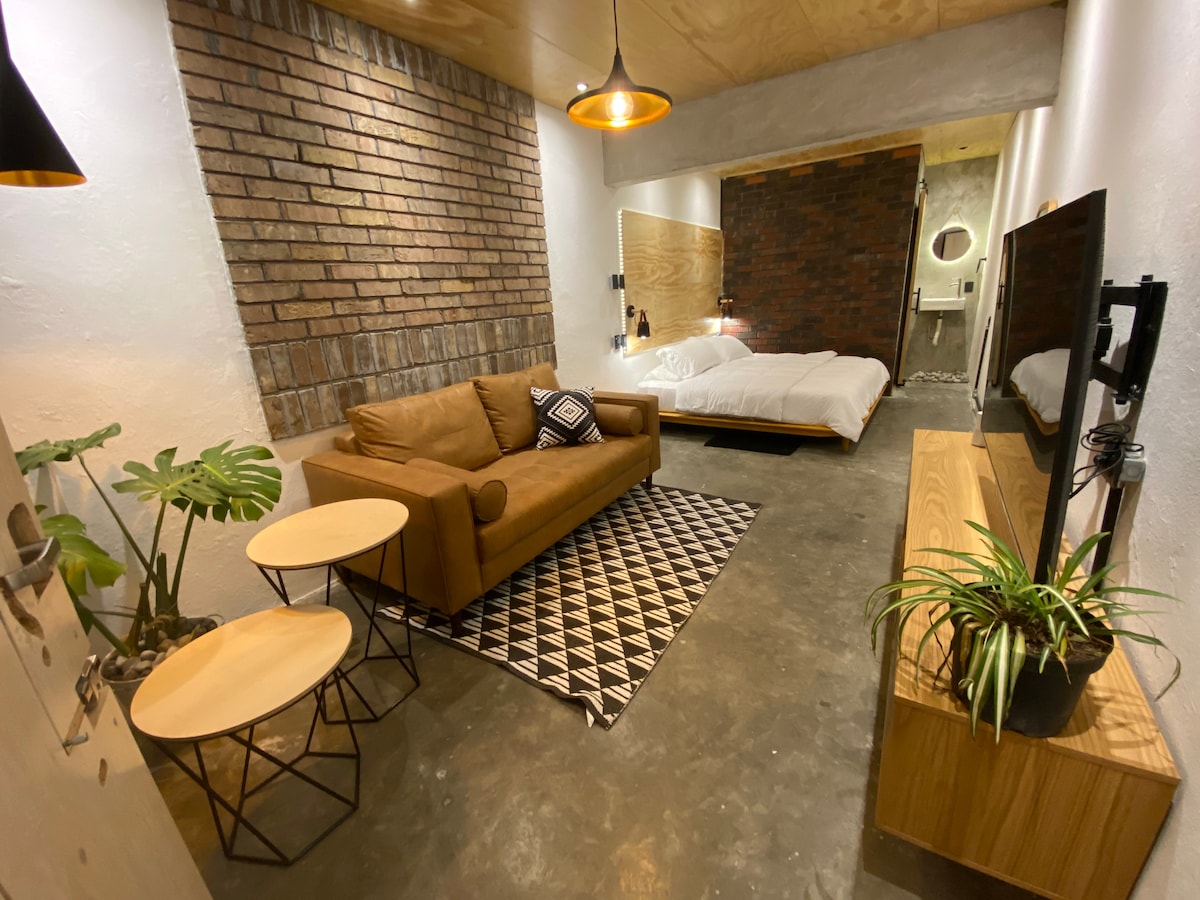 A studio space features a leather sofa positioned in front of a wall-mounted TV. Light wood cabinetry is placed beside the TV, with two small tables nearby. A king bed with white linens is located against a textured brick wall, enhancing the rustic atmosphere.
