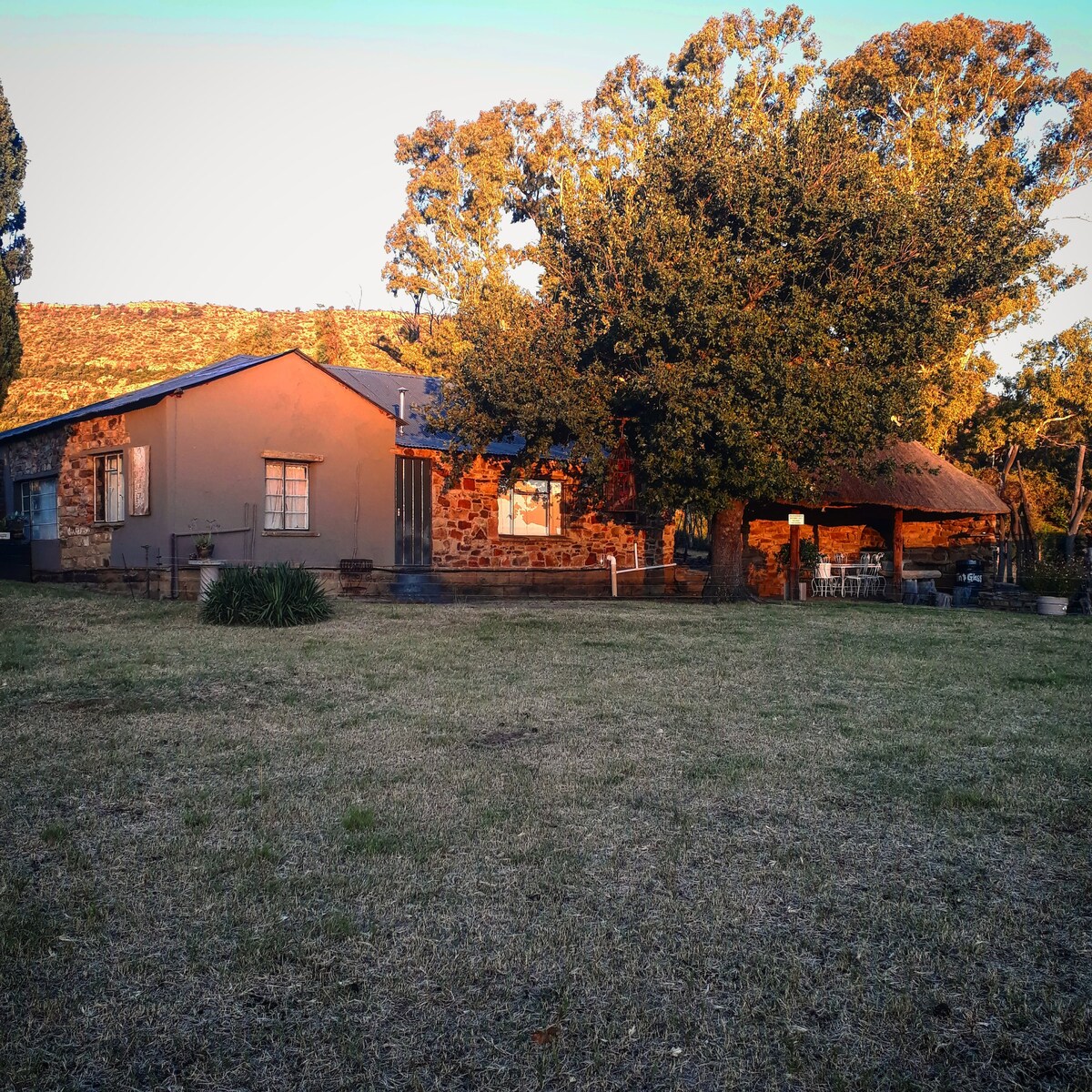 The stone cottage is set among a spacious lawn, featuring a blend of textured stone and painted exterior walls. A thatched roof extends over part of the structure. Tall trees provide shade, enhancing the serene rural landscape.