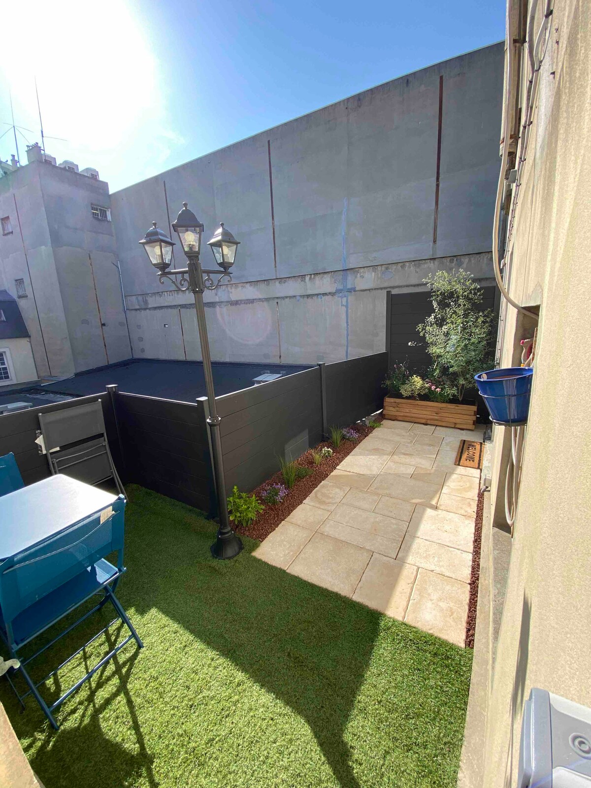 A private outdoor space features a well-maintained garden with artificial grass and stone pathways. Two blue chairs and a table are positioned in one corner, while decorative plants enhance the serene setting. A light fixture stands tall, providing illumination in the peaceful area.