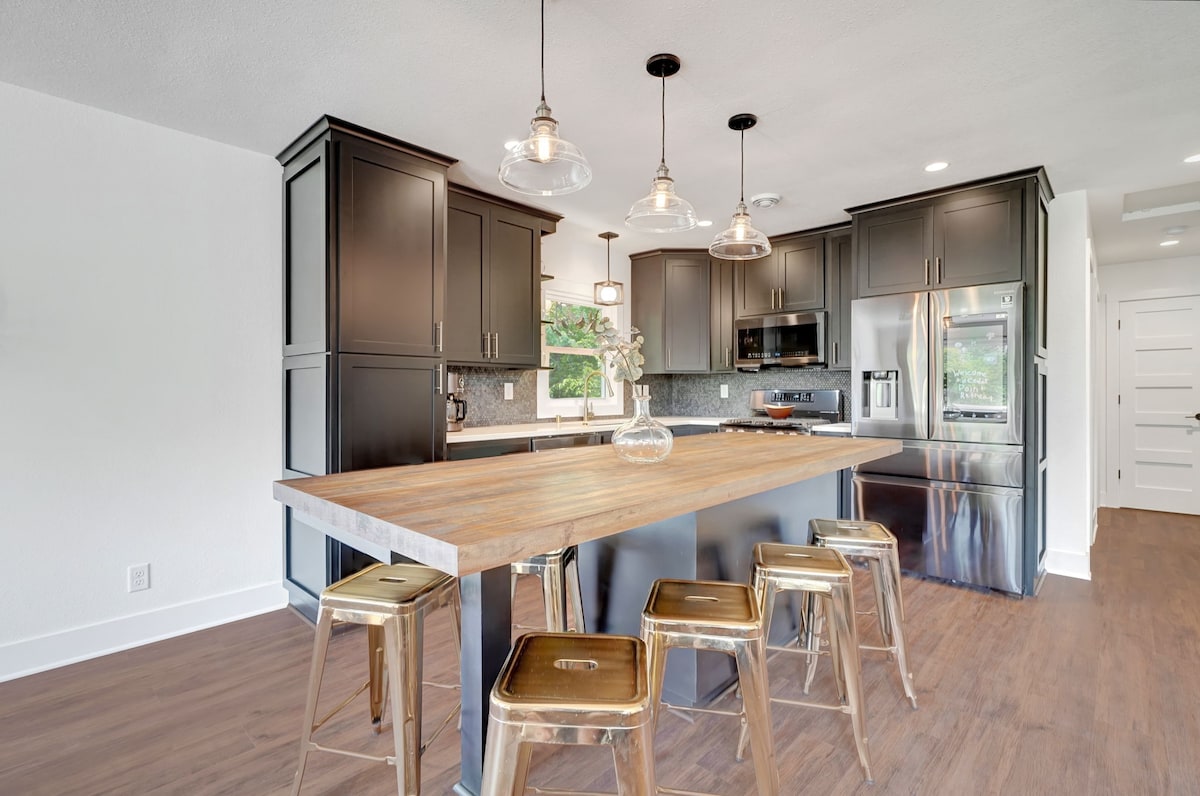 A modern kitchen is featured with sleek black cabinetry and a spacious wooden island. Four gold stools are positioned around the island. Natural light illuminates the area through a large window, highlighting the contemporary design and equipped appliances.