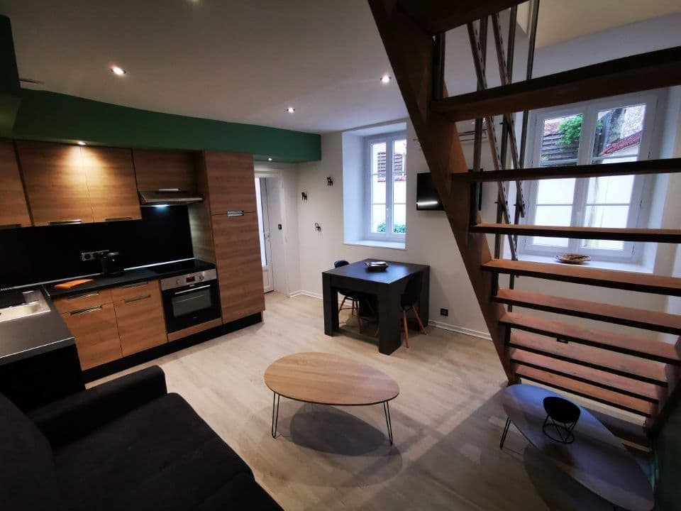 An open living space combines a modern kitchen with wood cabinetry and stainless-steel appliances. A dining area with a small table is positioned beside a seating area featuring a dark sofa. Natural light is provided by two windows, and a wooden staircase leads to the upper level.