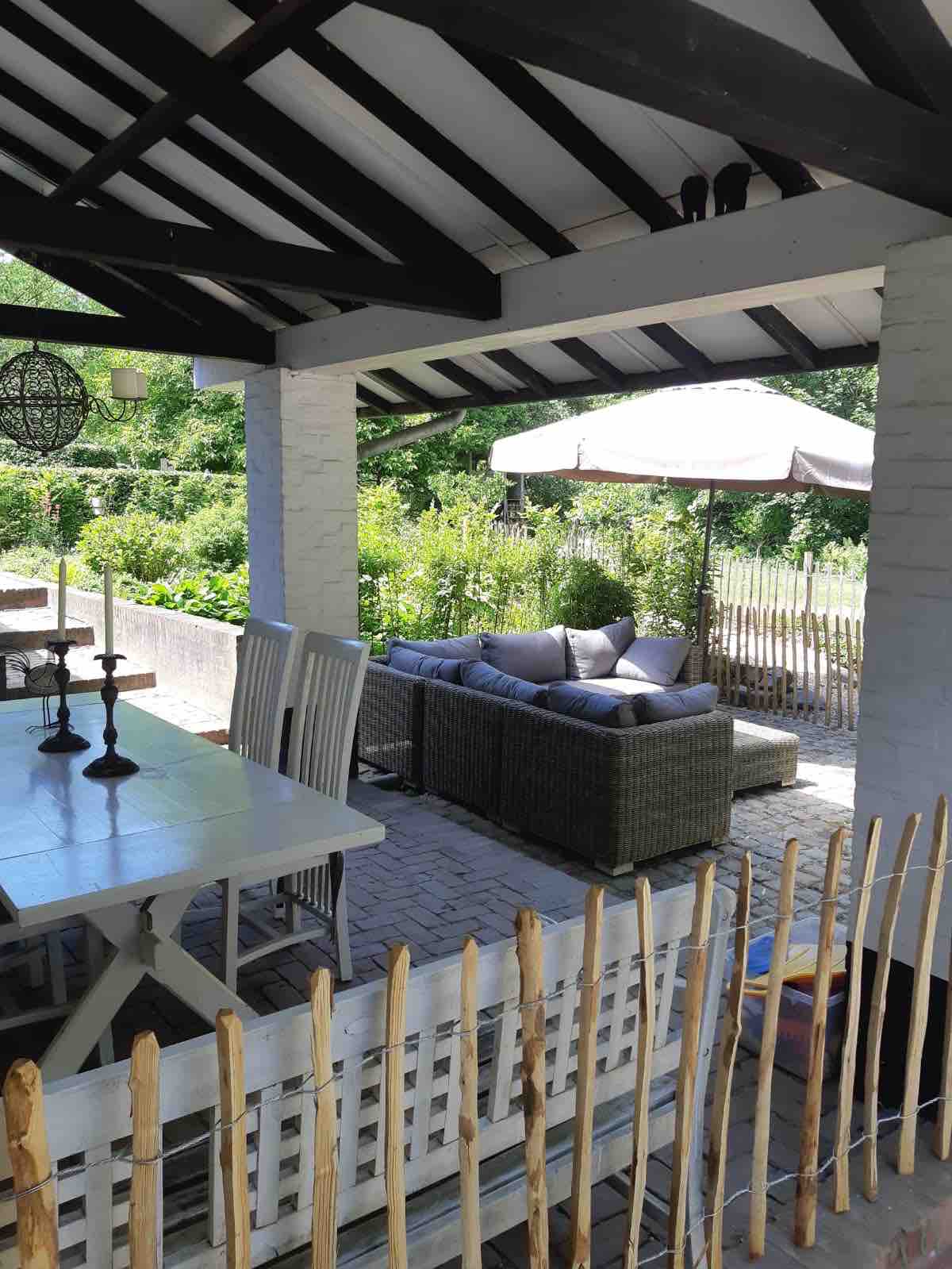 A shaded outdoor seating area features a comfortable woven sofa adorned with neutral cushions, surrounded by lush greenery. A dining table with several chairs is positioned nearby, and an umbrella offers additional shelter from the sun.