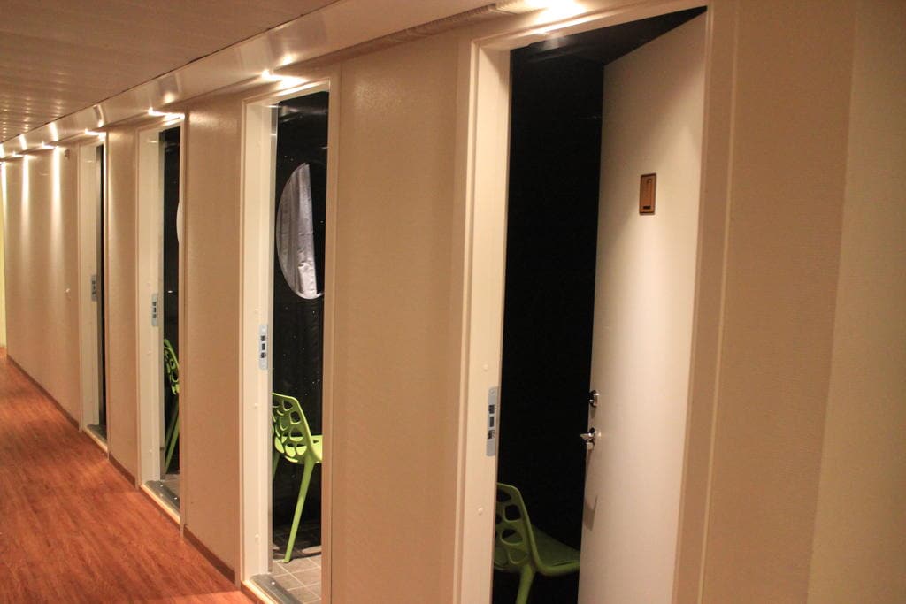 A corridor features several streamlined doors leading to private rooms. Each door is equipped with a secure lock and a small number is visible. Green chairs are positioned outside the rooms, providing a relaxed seating area.
