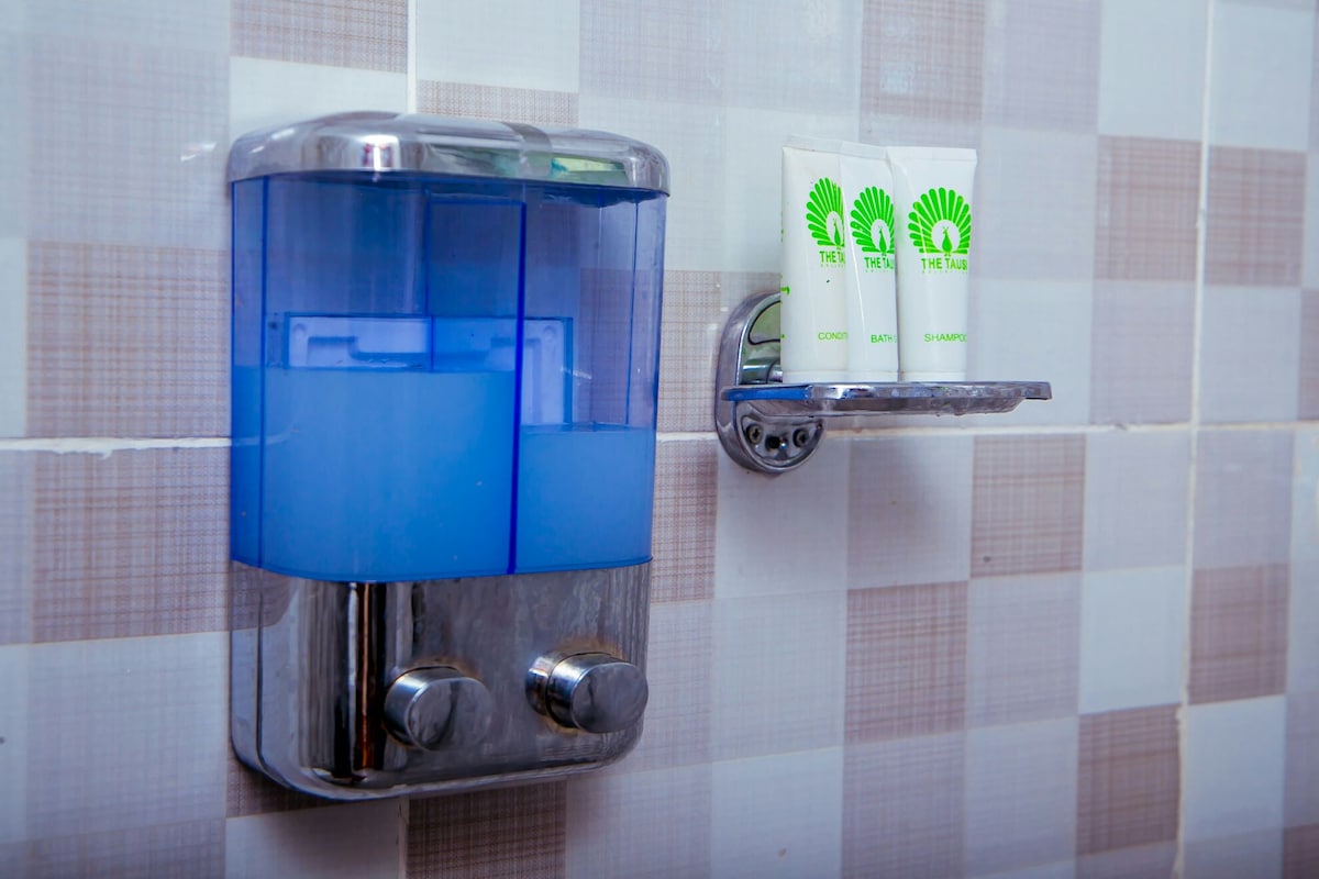 A wall-mounted soap dispenser in a clear blue container is paired with a metal shelf holding tubes of shampoo. The tiles feature a subtle checkerboard pattern in light beige and gray, enhancing the clean and functional aesthetic of the bathroom space.
