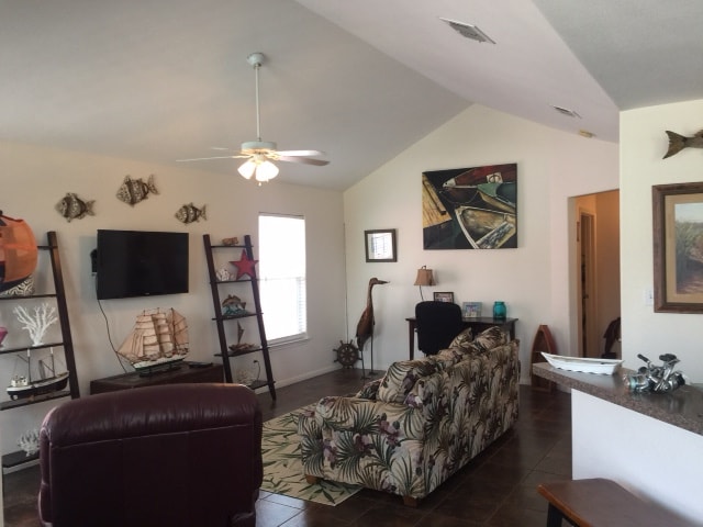 An open living room features a ceiling fan and large windows allowing natural light. A patterned couch is positioned near a TV, with tropical decorations and wall art enhancing the space. Access to a nearby hallway leads to additional areas of the home.