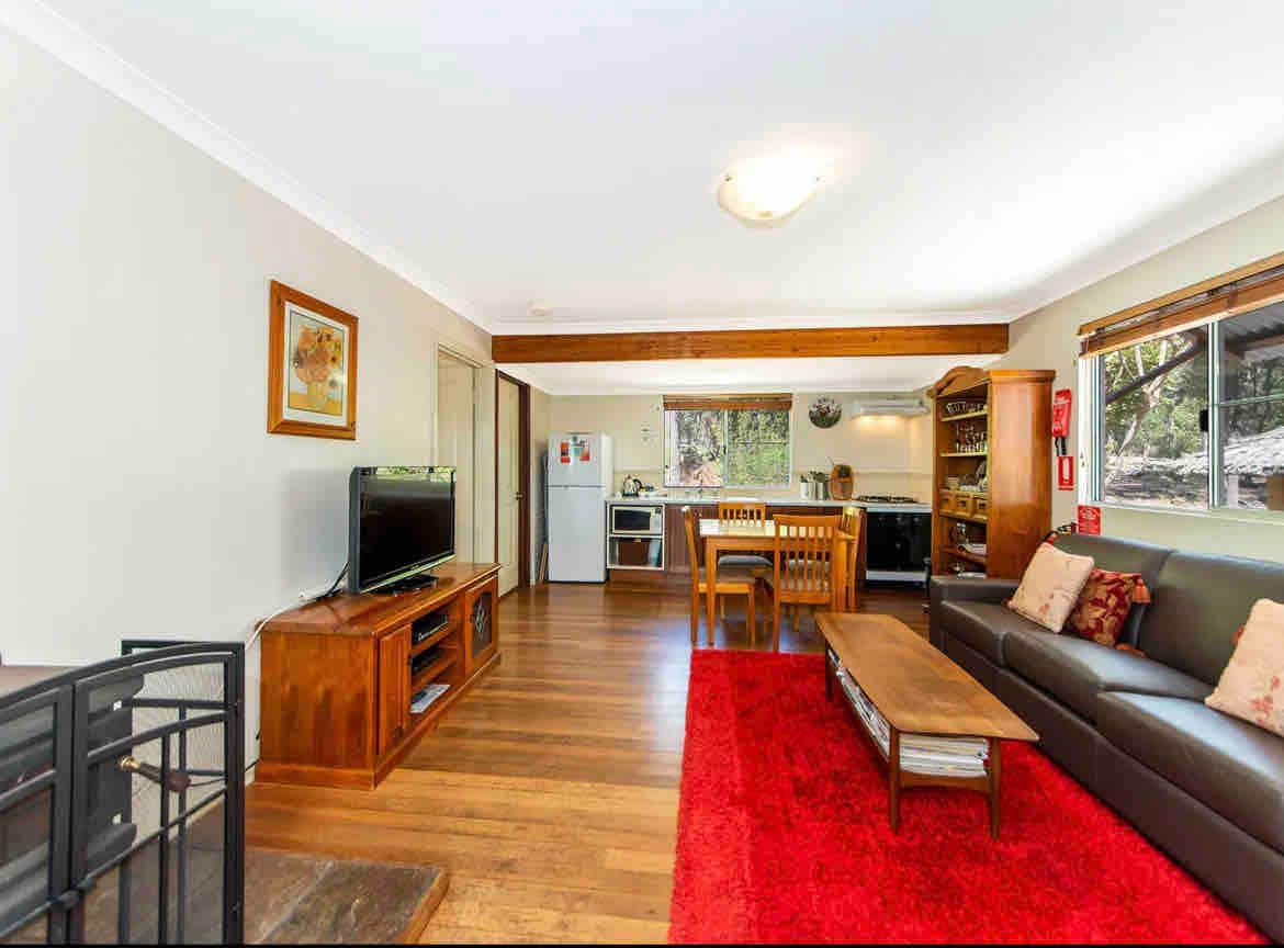 A comfortable living space is presented with a wooden dining table, seating four, positioned near a large window. A sofa sits on a vibrant red rug, alongside a wooden entertainment unit housing a television. The kitchen area is visible in the background, well-equipped with modern appliances.