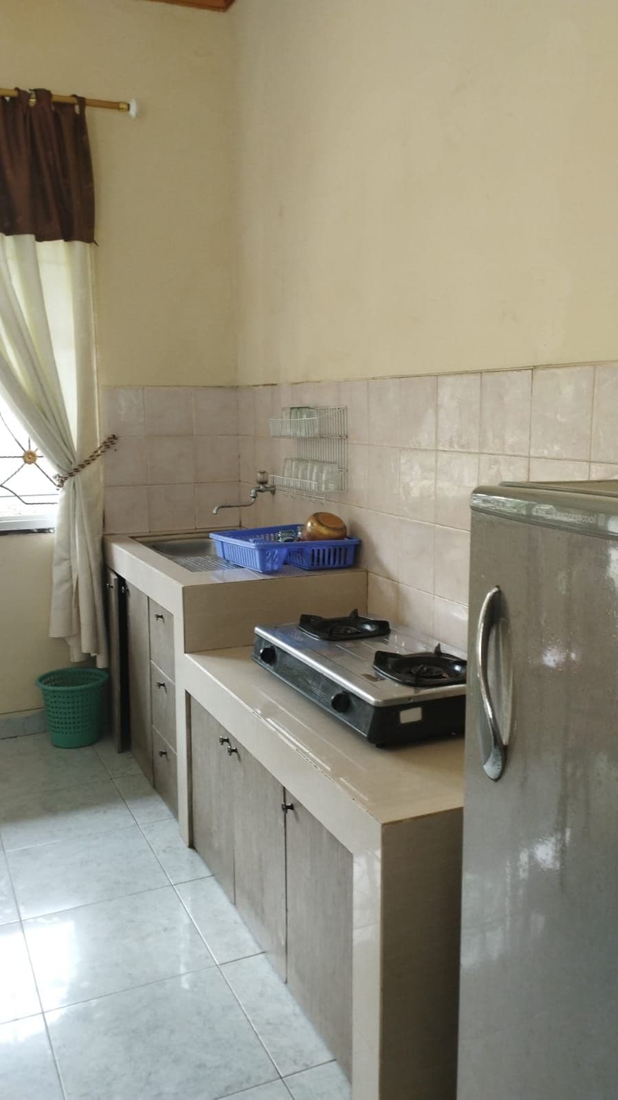 A functional kitchen area is presented, featuring light-colored cabinetry and a stainless steel refrigerator. A gas stove is positioned on the counter beside a sink, with a basket for utensils mounted above. Natural light enters through a window adorned with soft curtains.
