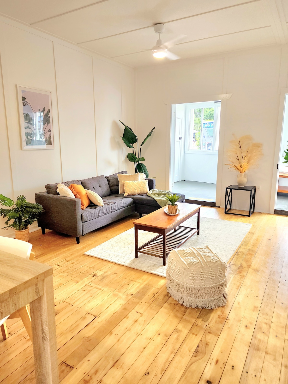 An open living area features a comfortable gray sofa adorned with decorative pillows. A wooden coffee table sits on a light rug, while natural light streams in through a nearby door. Green plants and a stylish decorative corner add warmth and character to the space.