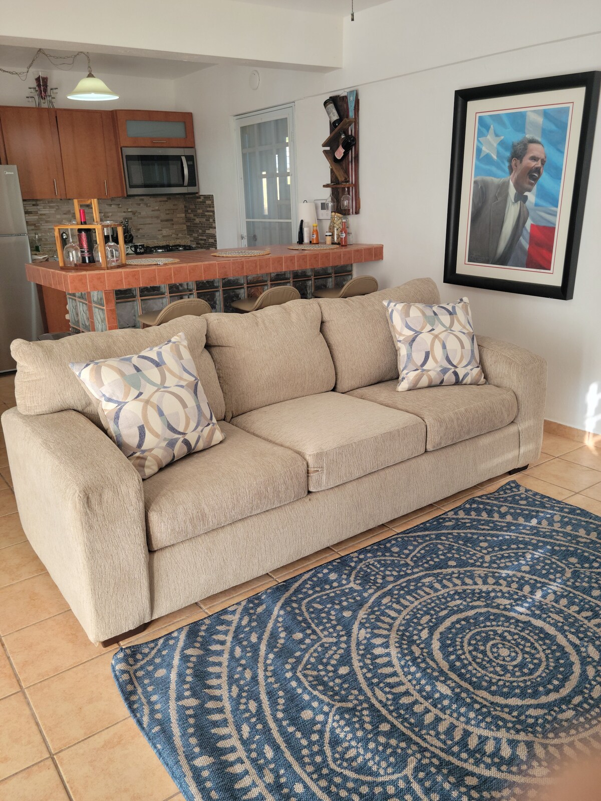 A spacious living area features a light-colored sofa with decorative pillows, positioned on a patterned area rug. A kitchen layout is visible in the background, showcasing wooden cabinetry and modern appliances. Natural light brightens the room through large windows, enhancing the inviting atmosphere.