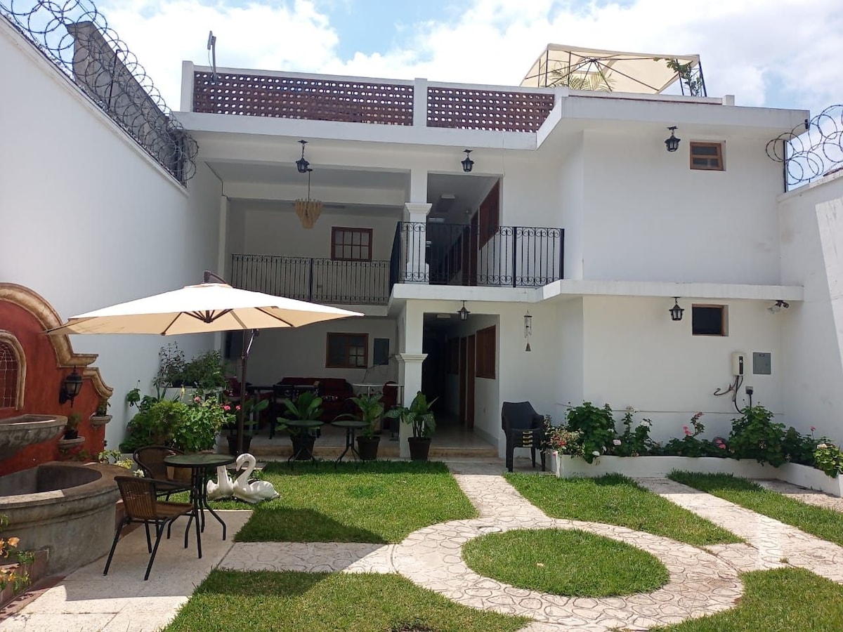 A multi-level property is surrounded by well-manicured gardens, featuring a seating area with an umbrella and decorative fountain. The upper level includes a terrace with railings, and various plants are scattered throughout, contributing to a serene outdoor atmosphere.