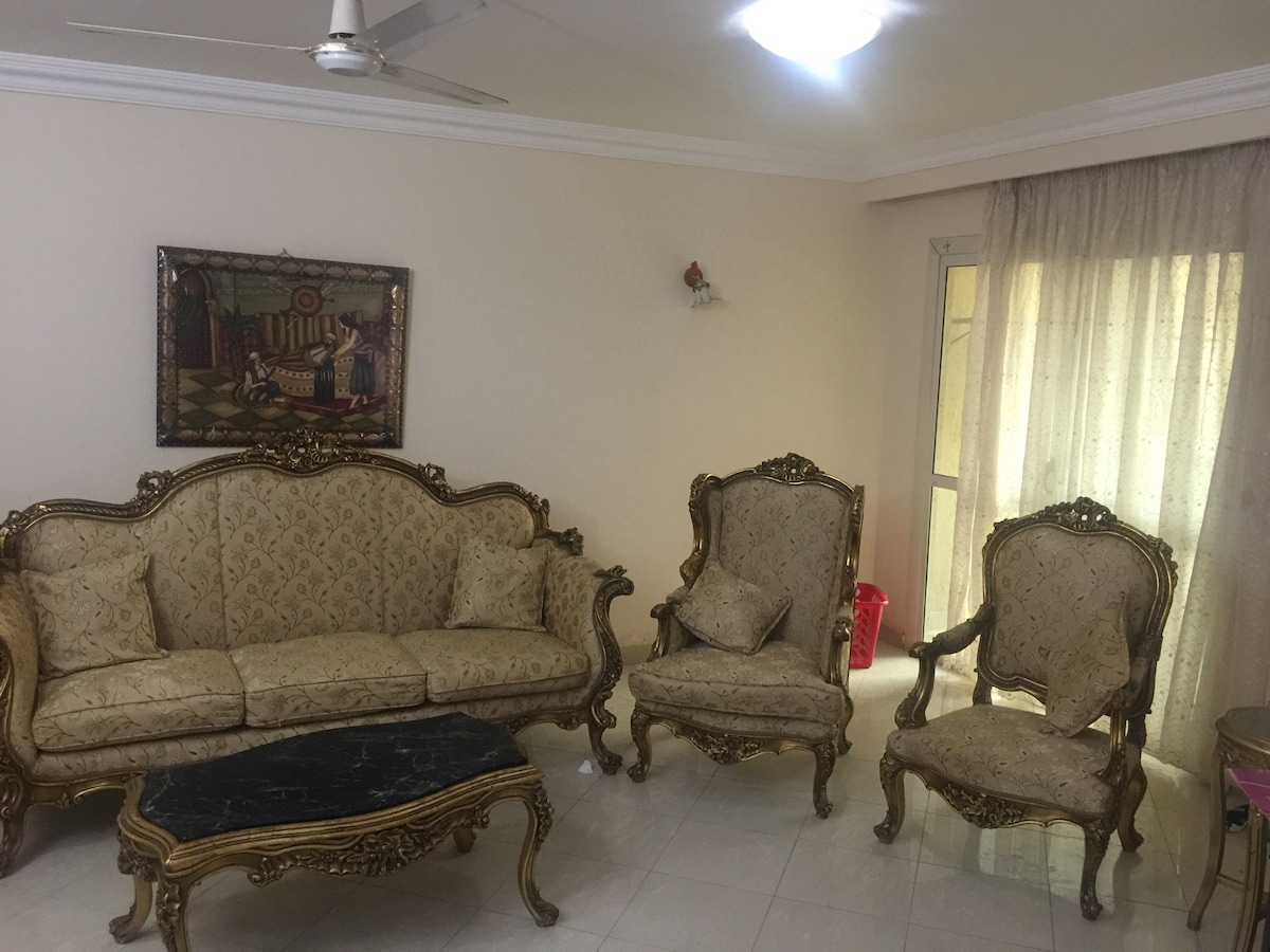 A well-furnished living room features an elegant seating arrangement with a decorative sofa and two matching chairs, all adorned with intricate gold detailing. A rectangular coffee table sits at the center, and a vibrant piece of wall art adds character to the space. Natural light filters through sheer curtains.