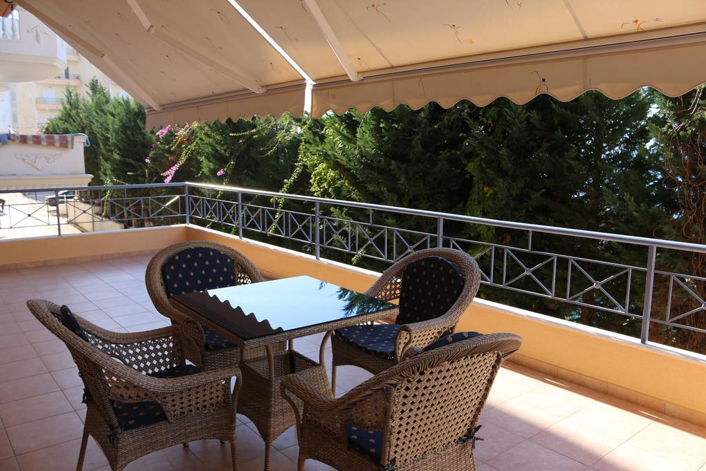 A covered terrace features a glass-top table surrounded by four woven chairs. The space is bordered by a railing and offers views of lush greenery, providing a serene spot for outdoor relaxation.