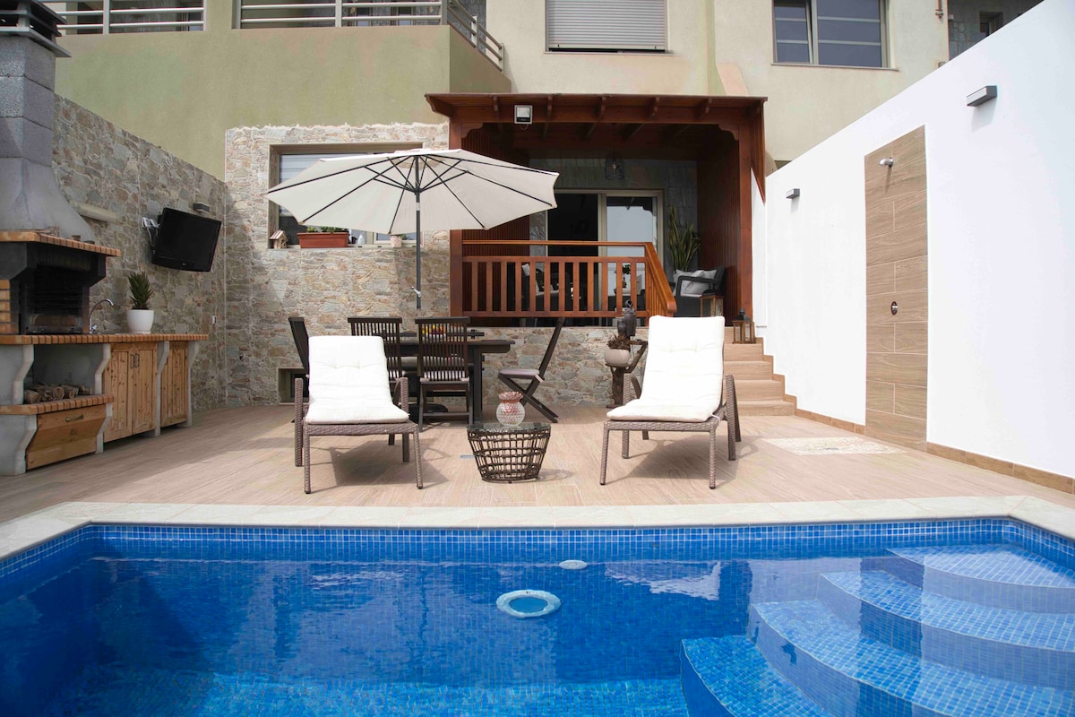 A private outdoor area features a warm-toned wooden deck surrounding a blue pool. Two lounge chairs are positioned beside the pool, while a large umbrella provides shade. A dining table with chairs is visible under a covered patio, and a built-in barbecue adds convenience to the space.