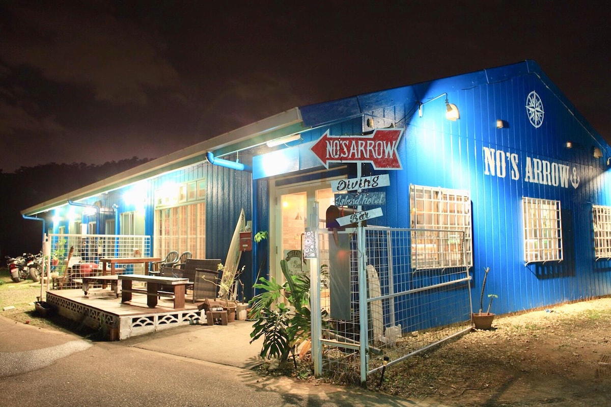 A blue building is illuminated under the night sky, featuring large windows and a welcoming entrance. Outdoor seating is arranged in front, with a wooden table and chairs visible. A sign indicates the establishment's name, adding character to the inviting exterior.
