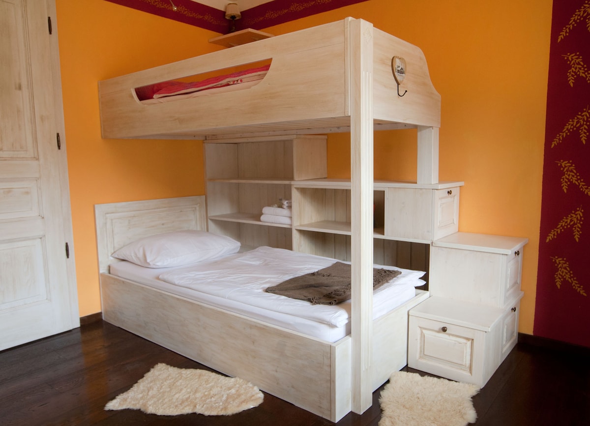 A wooden bunk bed is featured, with a spacious lower sleeping area and a higher sleeping area accessed by a small staircase. Shelves and a bedside table are incorporated into the design. Soft, neutral bedding contrasts with the vibrant orange walls.