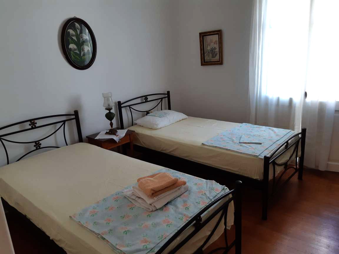 The bedroom features two single beds, each topped with light linens and decorative coverlets. A bedside table with a lamp is positioned between the beds. Natural light enters through sheer curtains, illuminating the simple decor, which includes a framed painting on the wall.