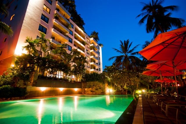The illuminated swimming pool reflects the night sky, surrounded by lush tropical vegetation. The multi-story building features balconies adorned with greenery, while vibrant red umbrellas provide shade over lounge chairs lining the poolside.