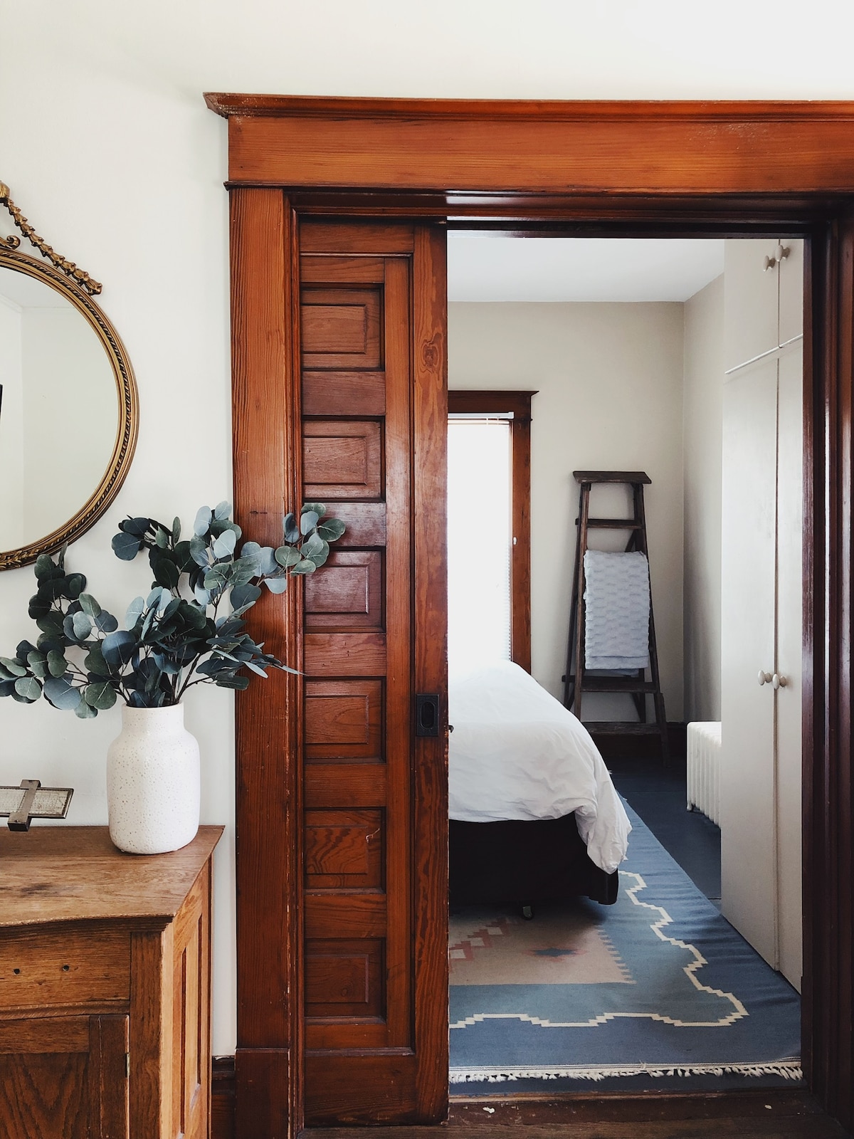 A doorway framed by rich woodwork leads into a bedroom. A spacious white bed is visible, alongside a light-colored wardrobe. A round mirror reflects natural light and a vase with greenery is placed on a rustic wooden cabinet in the foreground.