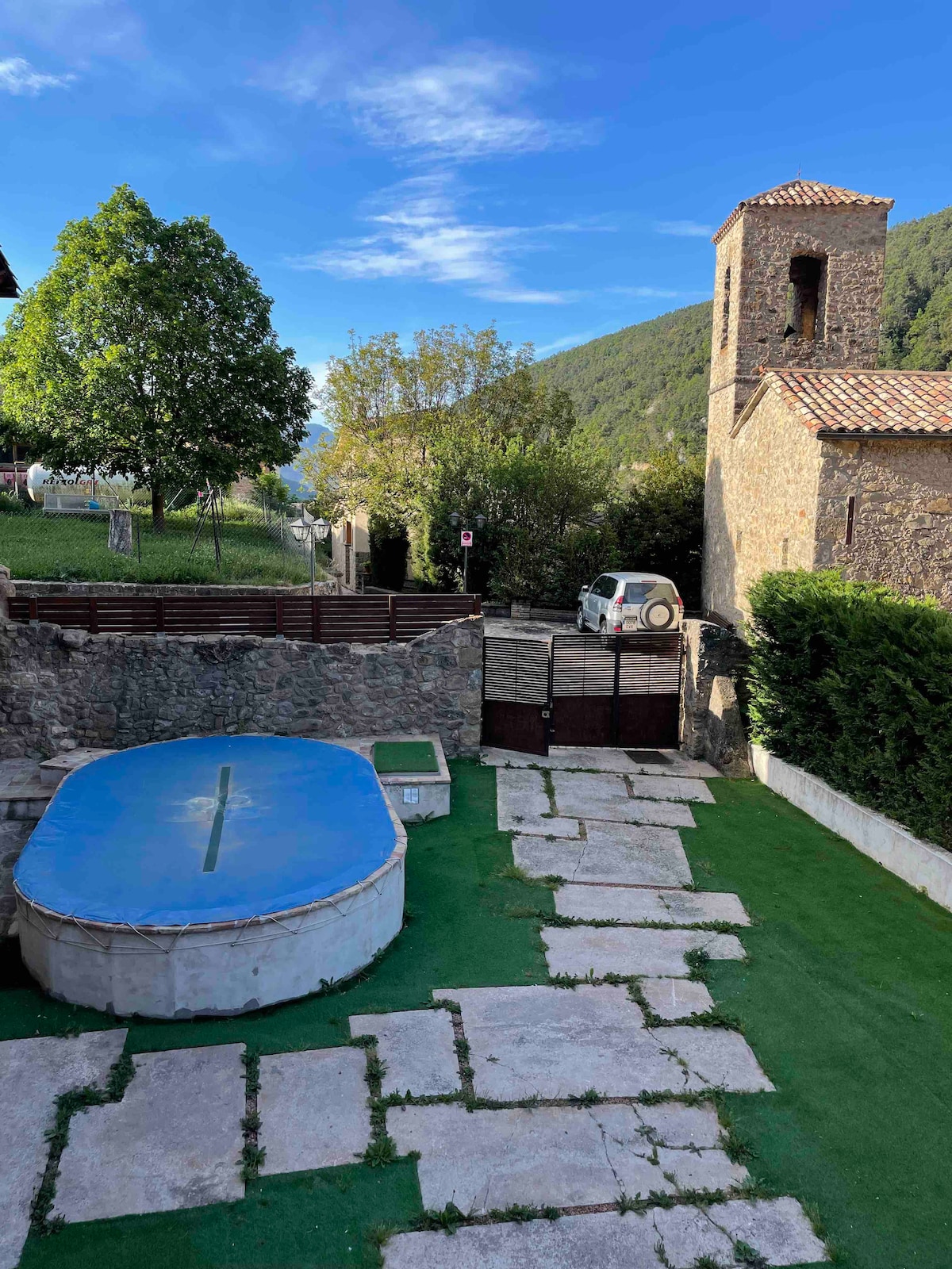 A tranquil outdoor area features a circular pool, bordered by manicured grass. Stone pathways lead to a gated entrance, with distant greenery and a towered structure set against a clear blue sky. Natural surroundings create a serene setting.