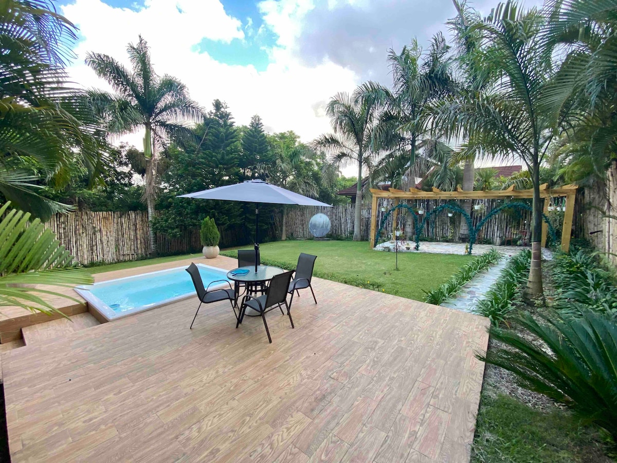 An outdoor space features a wooden deck with a table and four chairs shaded by an umbrella. A small pool is visible nearby, surrounded by manicured grass and tropical palm trees. A garden area includes a trellis and stone pathway leading to a well-kept lawn.