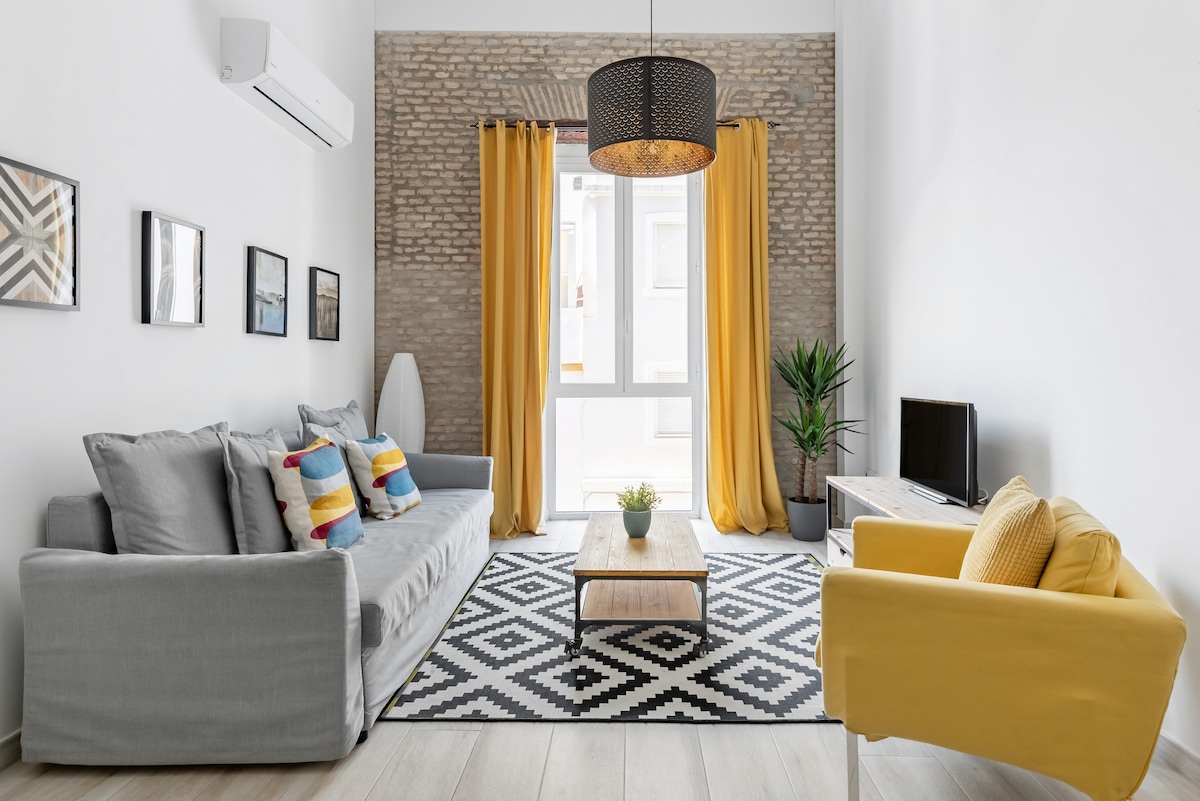 A bright living area features a gray sofa and a yellow accent chair. Colorful cushions are arranged on the sofa, and a patterned black and white rug covers the floor. Natural light enters through large glass doors, complemented by long yellow curtains and indoor plants.
