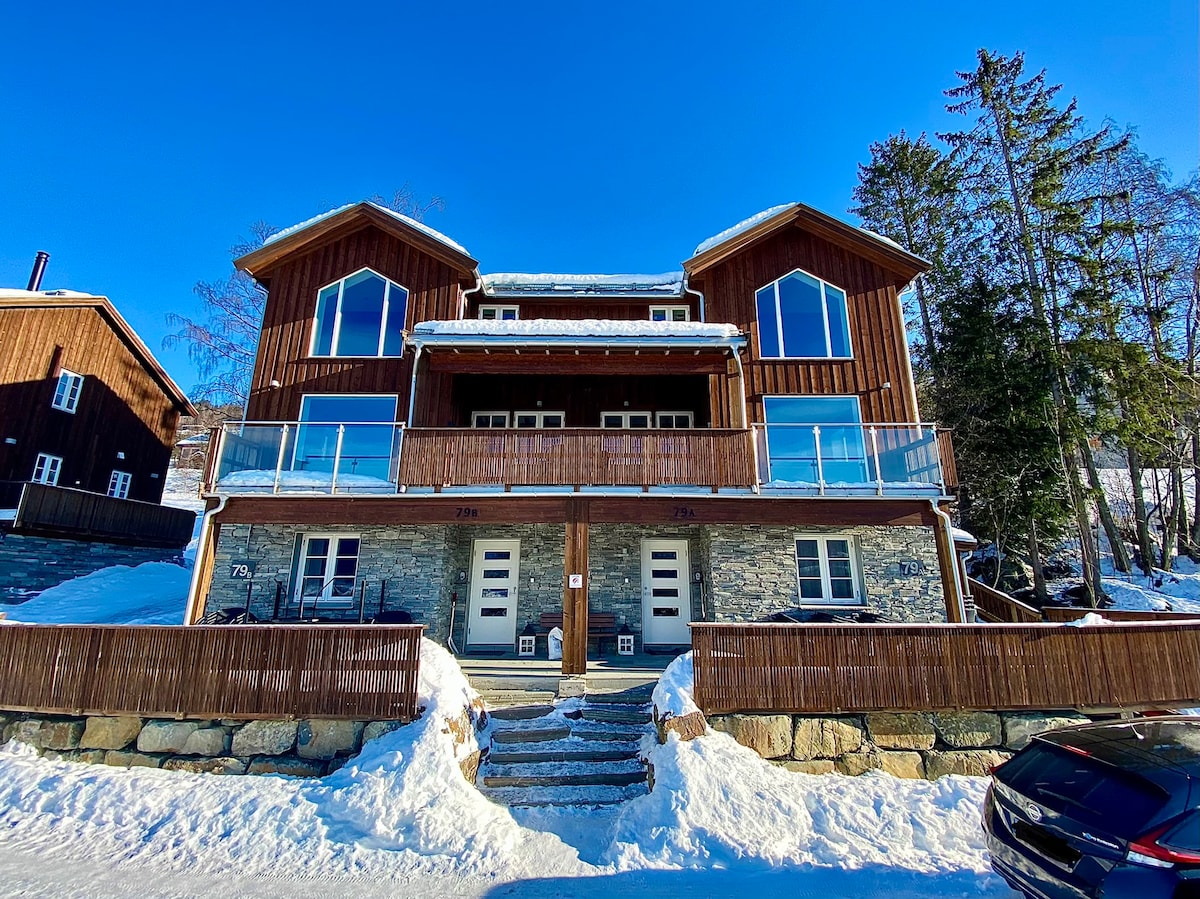 A twin cabin with a wooden exterior features large front windows and a balcony area. Steps lead up to the front entrance, framed by snow-covered ground. Surrounding trees provide a natural backdrop against the clear blue sky.