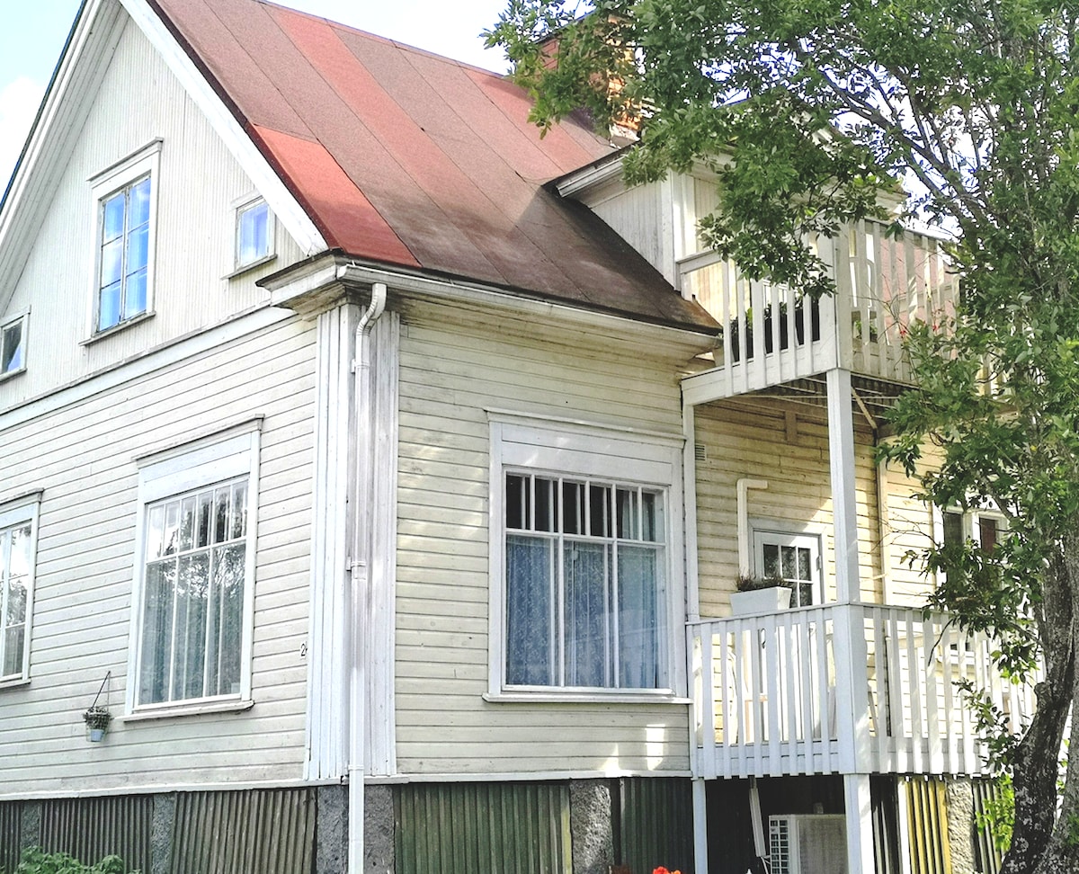 The exterior of a wooden house features a light-colored facade with large windows. A red sloped roof adds character, while a small balcony is visible on the upper level. Green foliage surrounds the property, contributing to the serene atmosphere.