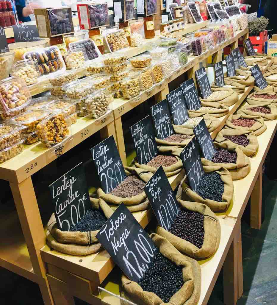 A vibrant market display features a variety of dried goods, including legumes and snacks, arranged in neat rows on wooden shelves. Each item is labeled with clear price signs, and burlap sacks hold different types of beans, showcasing an inviting selection of local culinary options.