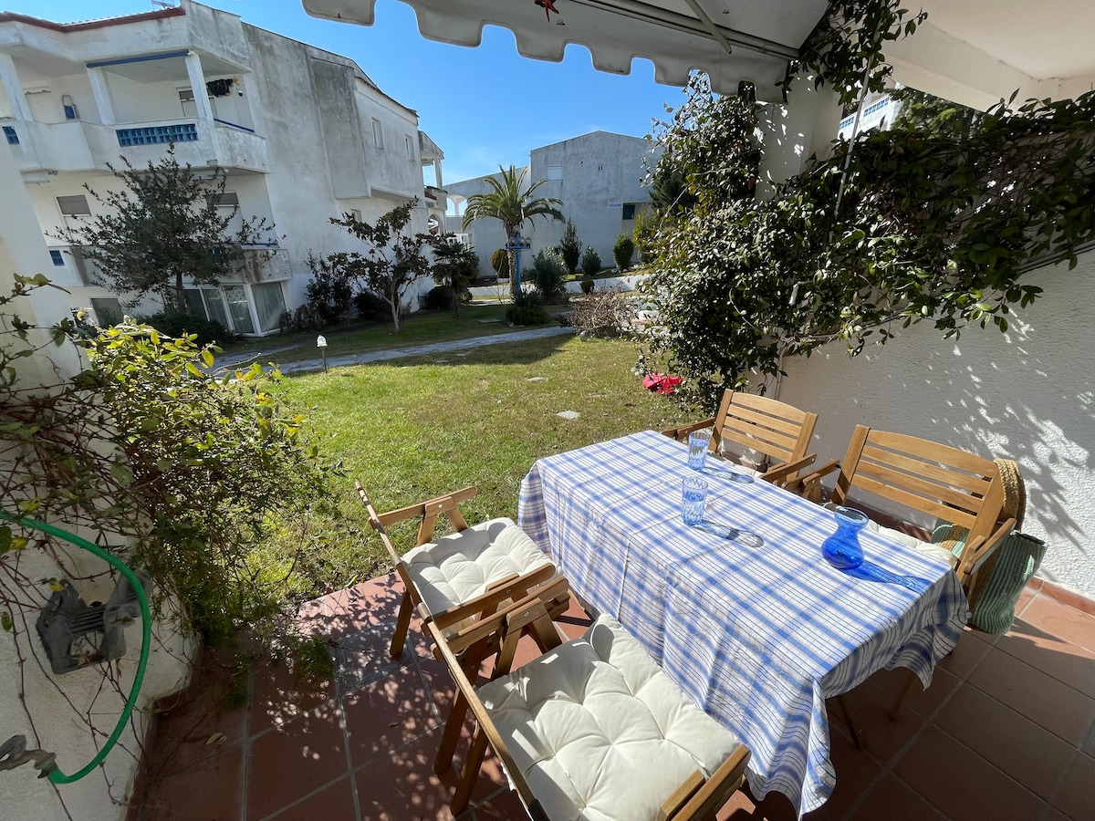 An outdoor dining area is set under a shaded awning, featuring a checkered tablecloth on a wooden table surrounded by four chairs. Lush greenery is visible in the garden, enhancing the space with natural surroundings, while sunlight pours in, creating a relaxed atmosphere.
