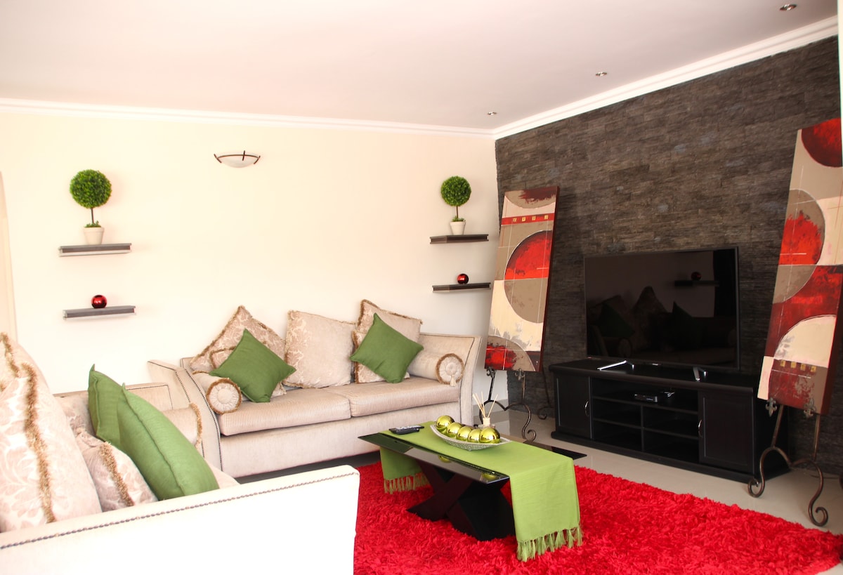 A stylish living area features a light-colored sofa adorned with green cushions and an array of decorative elements. A vibrant red rug lies under a modern black coffee table, which is decorated with green apples. A flat-screen TV is mounted against a textured wall.