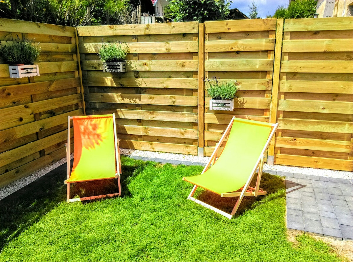 A private garden is presented with well-maintained grass, highlighted by two brightly colored lounge chairs. Wooden fencing creates a cozy ambiance, accented by potted plants that add greenery to the space. The area is designed for relaxation, bordered by a stone pathway.