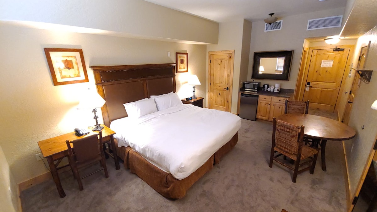 The studio accommodation features a King-sized bed with crisp linens, accompanied by bedside tables with lamps. A small round breakfast table for two is present, along with a kitchenette area equipped with a microwave and mini-fridge. A warm, neutral color scheme and wooden accents create a cozy environment.