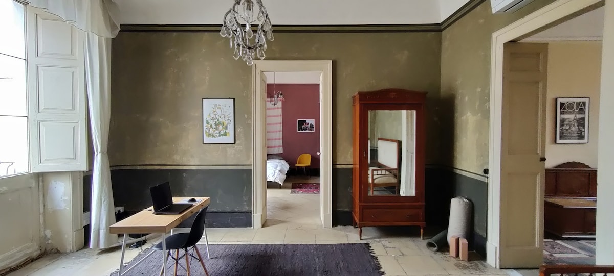 A spacious living area is highlighted by a decorative chandelier and contains a wooden desk with a chair. The walls display a muted color palette, and a mirror reflects the adjacent room. Natural light filters through large, shuttered windows.