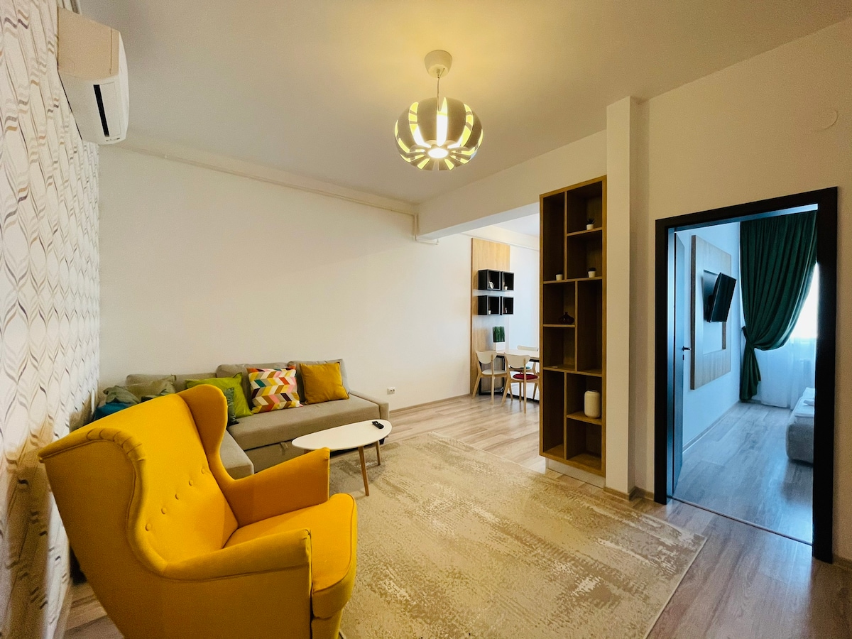 A living area is depicted with a bright yellow armchair and a light-colored rug. A grey sofa accompanies a modern coffee table, while a shelving unit adds storage and display space. Natural light enters from a doorway leading to another room, featuring green curtains.