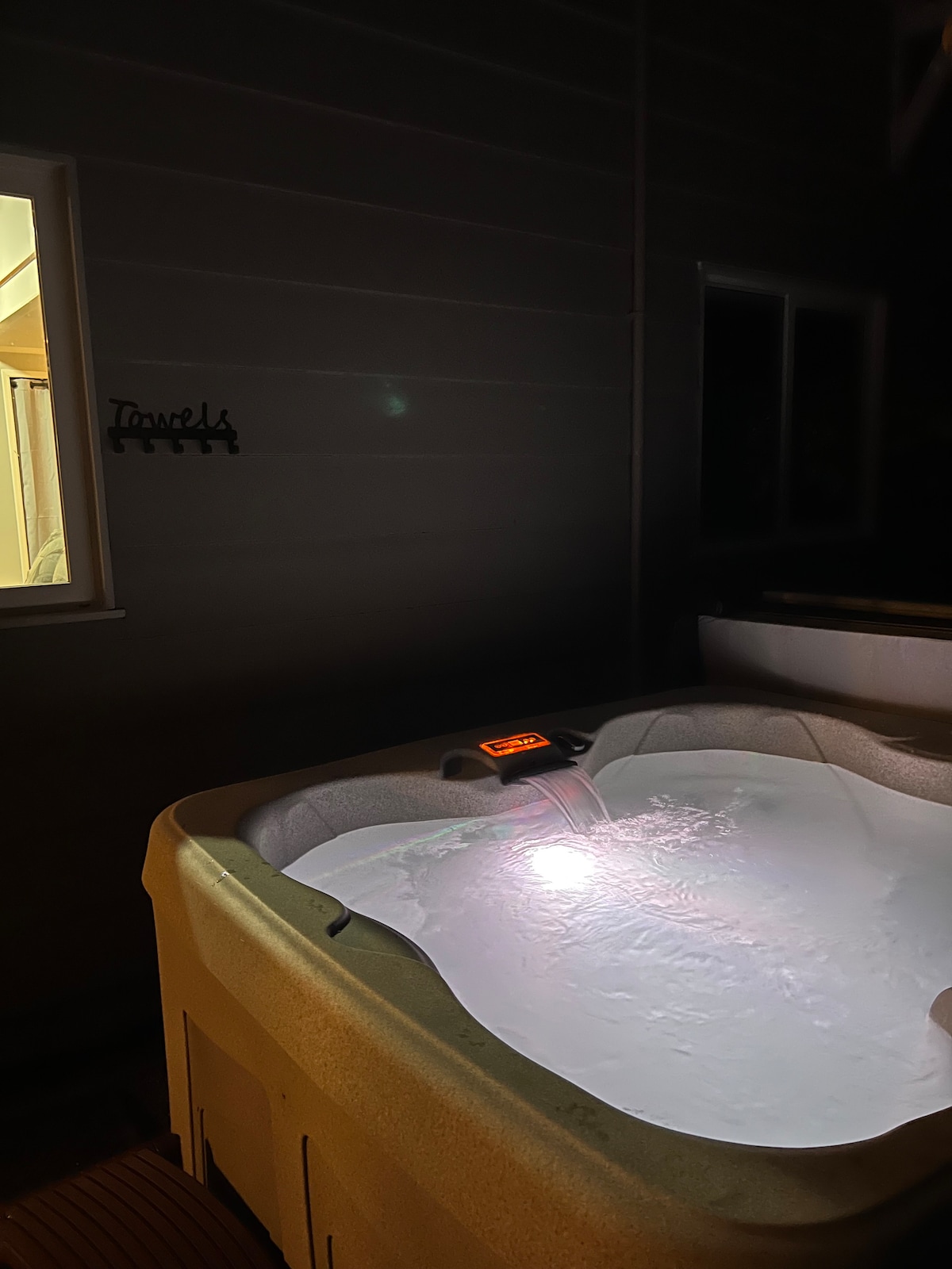 A hot tub is illuminated gently, showcasing clear water rippling under soft lighting. The surrounding area features a window that reveals a glimpse of indoor space, contributing to a cozy and private atmosphere.