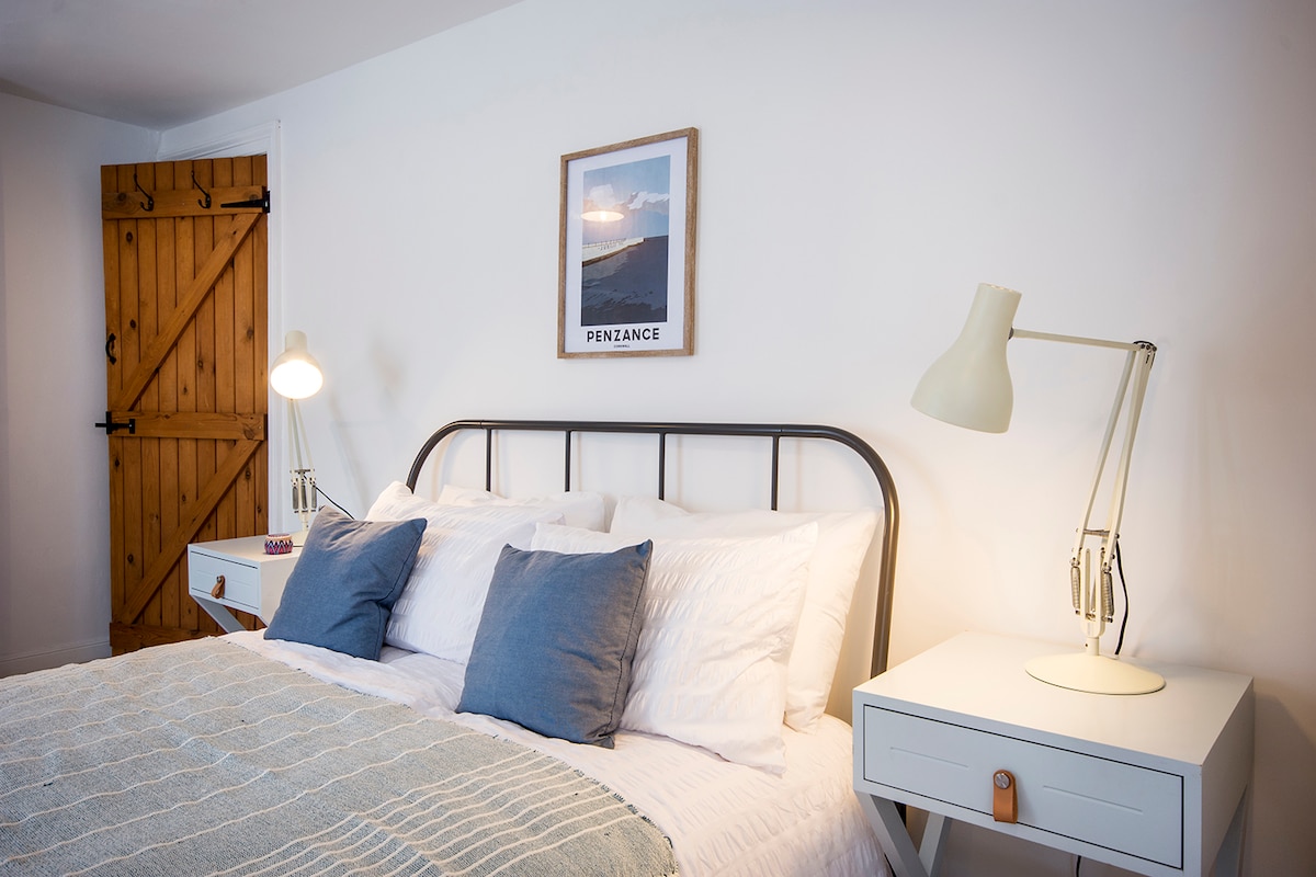 A cozy bedroom features a spacious bed with soft white linens and decorative blue pillows. A framed print titled 'Penzance' hangs above the bed. Bedside tables with lamps are positioned on either side, and a wooden door adds a rustic touch to the space.