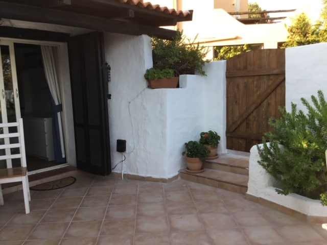 A private patio area is presented with terracotta tiles and a wooden gate. Potted plants are arranged along the walls, adding greenery. A small table and chair are visible, providing a cozy spot for relaxation. The entrance to the studio is framed by white walls and a dark door.