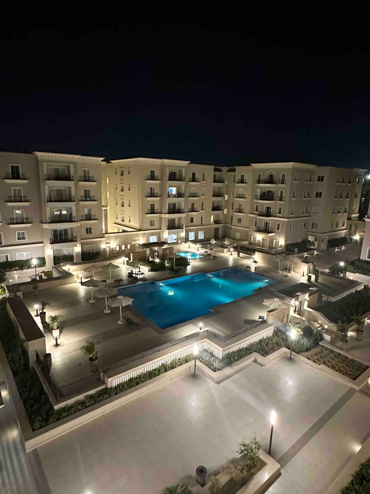 An illuminated outdoor area is visible at night, featuring a large swimming pool surrounded by lounge chairs and well-maintained gardens. The surrounding buildings are softly lit, creating a welcoming ambiance. Pathways lead to the pool, enhancing accessibility.