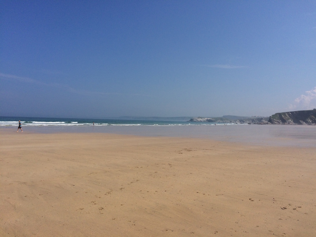 A wide sandy beach stretches under a clear blue sky, with gentle waves lapping at the shore. The scene captures the expanse of golden sand meeting the ocean, ideal for leisurely strolls or relaxing by the water.