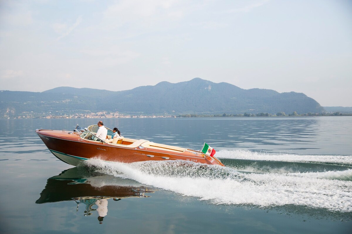 A sleek wooden boat glides across the calm water, creating gentle waves. The image captures two passengers enjoying the ride, with scenic mountains visible in the background and a hint of greenery along the shoreline.