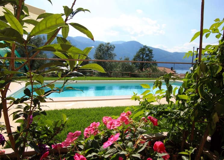 An outdoor area features a swimming pool surrounded by lush greenery and vibrant flowers. The pool reflects the sky and mountains in the background, creating a serene atmosphere. Sun loungers are positioned near the pool, inviting relaxation.