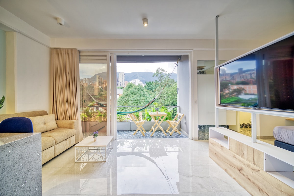 A spacious living area features a comfortable sofa facing a large flat-screen TV. Natural light fills the room from expansive glass doors that open to a balcony with a hammock and a small outdoor dining set, providing a view of the lush surroundings.