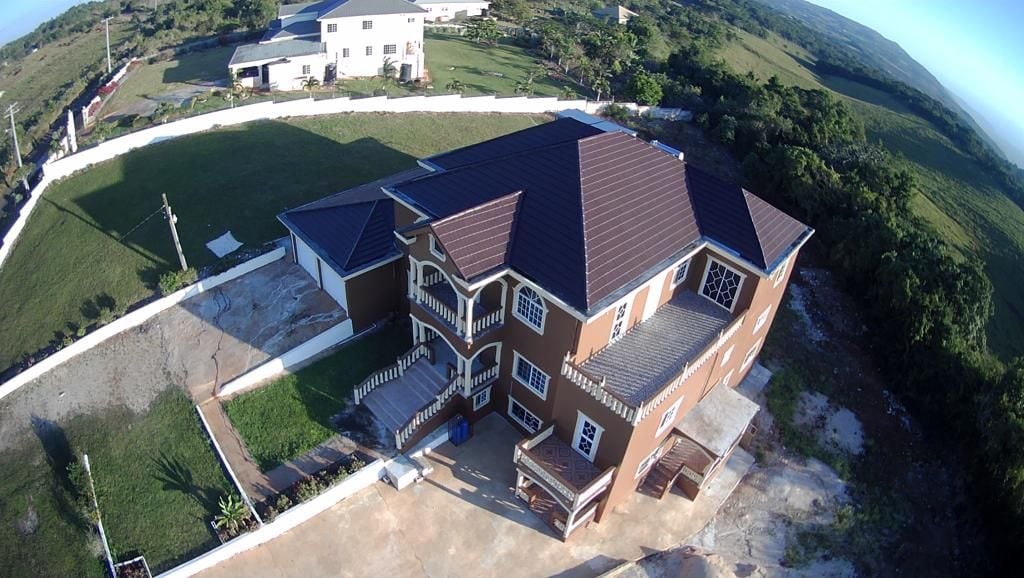 The aerial view captures a spacious multi-story house with a brown exterior and multiple balconies. Surrounding green fields and rolling hills are visible, along with a protective white fence enclosing the property. Nearby structures can be seen in the background, providing additional context.