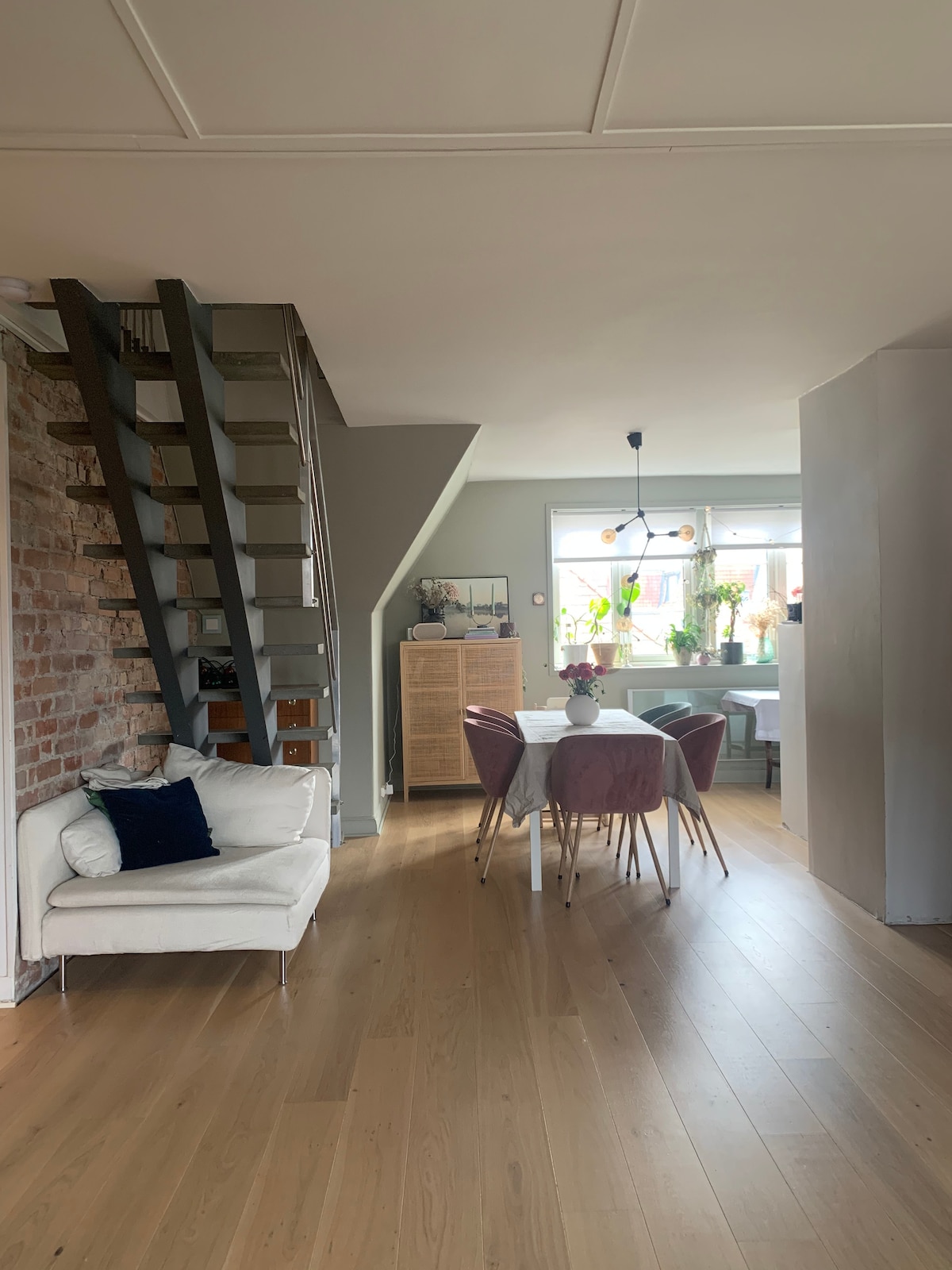 A bright and airy living space features a mix of natural wood flooring and exposed brick walls. A small white sofa is positioned next to a modern dining table surrounded by four upholstered chairs. Natural light enters through large windows adorned with potted plants.