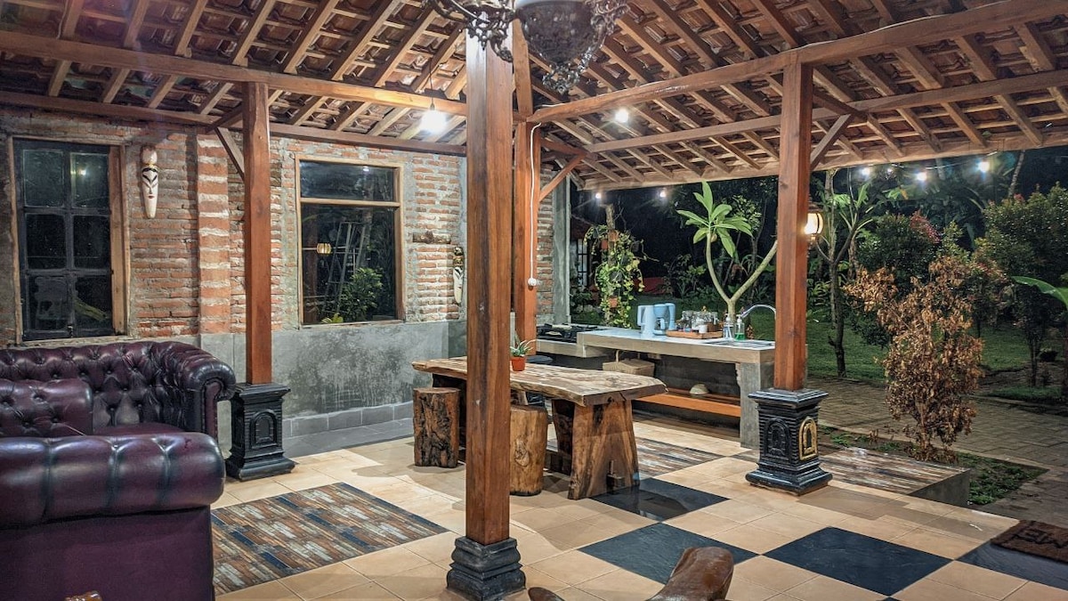 A covered outdoor lounge area features wooden beams and a classic brick wall. A large, dark leather sofa and wooden tables provide seating and dining space. Lush greenery can be seen through the windows, while warm lighting enhances the inviting atmosphere.